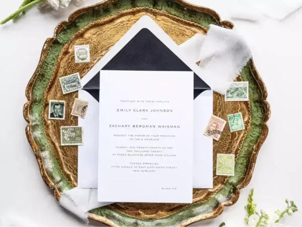 A wedding invitation on a white card placed on a gold and green plate, with a white envelope behind it. Vintage stamps are scattered around the invitation and envelope.